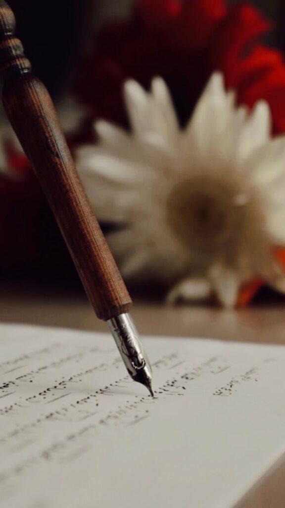 Close-up of a wooden fountain pen writing calligraphy on paper with blurred flowers in the background.